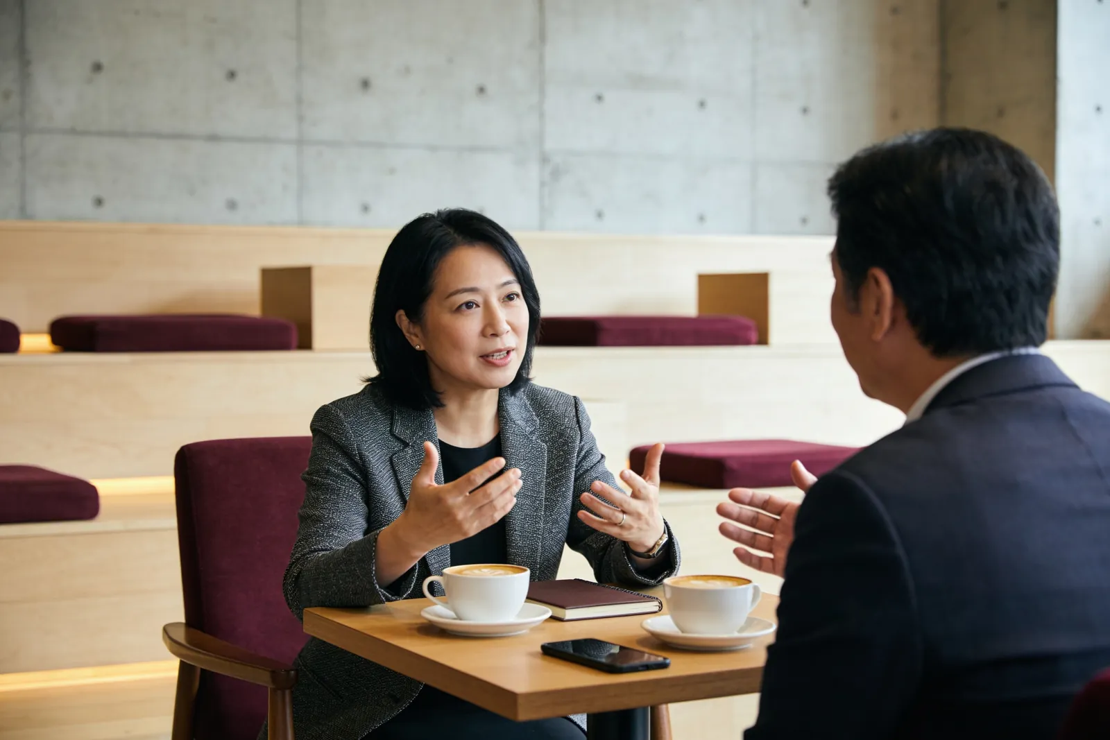 咖啡館裡兩位成員面對面坐著，女士一邊比手勢一邊說明，男士在聽，桌上兩杯咖啡和一本筆記本。